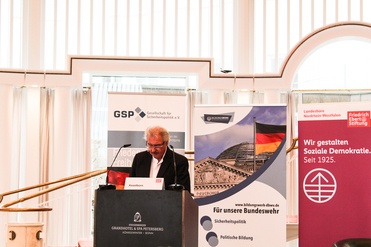 Jean Asselborn spricht an einem Rednerpult vor Bannern der GSP, des Bildungswerks Bundeswehr und der Friedrich-Ebert-Stiftung.