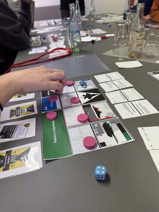 Eine Hand bewegt pinke Spielsteine auf einem Strategie-Brettspiel. Spielkarten, ein blauer Würfel und Getränkeflaschen sind auf dem Tisch.