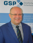Lächelnder Mann mit Brille und blauem Sakko vor einem Banner der GSP (Gesellschaft für Sicherheitspolitik).