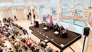 Podiumsdiskussion mit fünf Männern auf der Bühne vor einem Publikum in einem hellen, modernen Veranstaltungssaal.