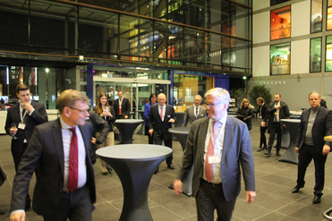 Geschäftsleute in Anzügen unterhalten sich auf einer Empfangsveranstaltung mit Stehtischen und moderner Glasarchitektur.