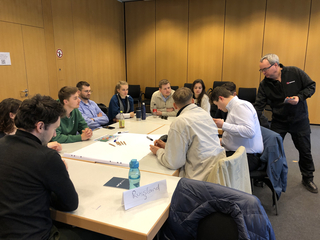Eine Gruppe junger Erwachsener sitzt an einem Tisch in einem Workshop, während ein Dozent daneben steht.