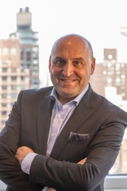 Lächelnder glatzköpfiger Mann im dunklen Anzug mit verschränkten Armen, vor einem Fenster mit Stadtblick.