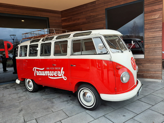 Vintage rot-weißer VW T1 Bus mit 'Traumwerk'-Schriftzug und Dachgepäckträger, vor modernem Gebäude geparkt.