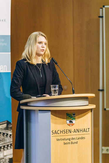 Eine blonde Frau in dunklem Anzug spricht an einem Rednerpult mit dem Wappen und Schriftzug von Sachsen-Anhalt.