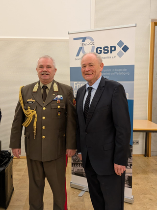 Zwei lächelnde Männer, einer in Militäruniform mit Orden, der andere im Anzug, vor einem GSP-Banner stehend.