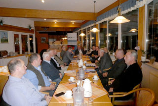 Männer sitzen bei einem Treffen an einem langen Tisch in einem Restaurant. Getränke auf dem Tisch, GSP-Banner im Hintergrund.
