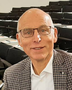 Lächelnder Mann mit Glatze und Brille in kariertem Sakko, sitzend in einem Hörsaal.