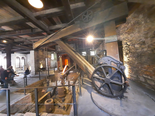 Historisches Industriegebäude-Interieur mit großen Holzbalken, alten Maschinen, hellem Ofenglühen und Besuchern.