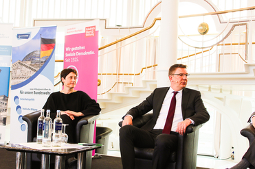 Zwei Sprecher, eine Frau und ein Mann, sitzen auf Sesseln bei einer Konferenz mit Bundeswehr- und FES-Bannern.