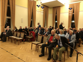 Zahlreiche Personen, meist ältere Männer, sitzen in einem Saal mit goldbraunen Vorhängen und Parkettboden, manche mit Getränken.