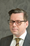 Professionelles Porträt eines Mannes mit dunklen Haaren und Brille, Anzug und gestreifter Krawatte, vor grauem Hintergrund.