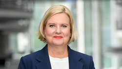 Nahaufnahme einer blonden Frau mit rotem Lippenstift und blauem Blazer, leicht lächelnd in die Kamera blickend.