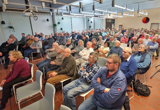 Große Gruppe von Erwachsenen und Senioren sitzt in einem hellen Saal und blickt aufmerksam nach vorne.