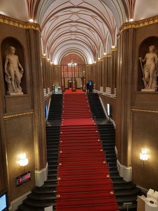 Eine große Halle mit einem langen roten Teppich auf einer breiten Treppe, flankiert von Säulen und Statuen, die zu einem gewölbten Eingang führt.