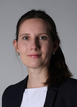 Porträt einer lächelnden Frau in dunkelblauem Blazer, weißem Shirt und Jeans vor hellem Hintergrund.