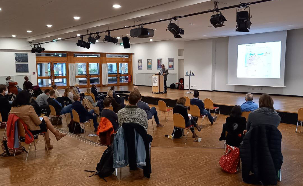 Eine Person hält einen Vortrag auf einer Bühne vor Publikum in einem großen Saal mit einer Karte auf der Projektionsfläche.