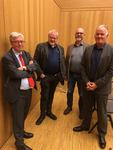 Vier lächelnde Männer in Geschäftskleidung stehen in einem holzvertäfelten Raum.