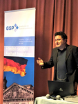Ein Mann präsentiert bei einem GSP-Event vor einem Banner mit Deutschlandflagge und Reichstag. Laptop und Mikrofone.