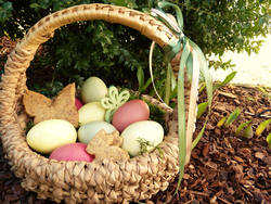 Ein Osterkorb aus Stroh, gefüllt mit bunten Eiern und Hasenkeksen, steht zwischen grünen Büschen und Rindenmulch.