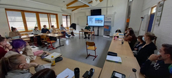 Schüler und Redner mit Masken bei einer Präsentation im Klassenzimmer mit Bildschirm.