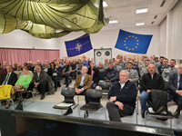 Zuschauer in einem Saal mit NATO- und EU-Flaggen und grünen Fallschirmen an der Decke. Einige Personen tragen Militäruniformen.