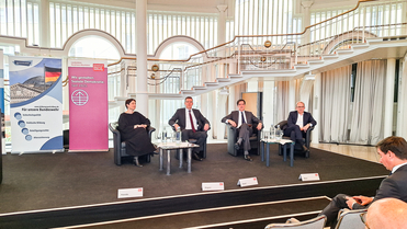 Vier Podiumsteilnehmer, drei Männer und eine Frau, sitzen in Sesseln auf einer Bühne bei einer Diskussion, im Hintergrund Konferenzbanner.