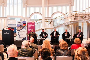 Podiumsdiskussion mit fünf Männern (einem Offizier) vor Publikum in einem hellen Saal mit Treppe und Bannern.