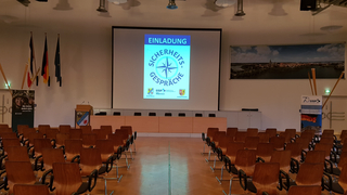 Leerer Konferenzraum mit Reihen von Holzstühlen, einem Rednerpult und einer Leinwand, die 'Sicherheits-Gespräche' anzeigt.