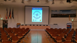 Leerer Konferenzraum mit Reihen von Holzstühlen, einem Rednerpult und einer Leinwand, die 'Sicherheits-Gespräche' anzeigt.