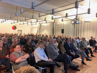Ein vielfältiges Publikum, hauptsächlich Männer, sitzt in Reihen und verfolgt aufmerksam eine Präsentation in einem hellen Saal mit Beamer.