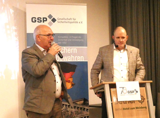 Zwei Männer auf einer GSP-Veranstaltung. Einer spricht ins Mikrofon, der andere steht am Rednerpult vor dem GSP-Logo.