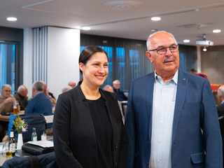 Eine lächelnde Frau und ein Mann mit Schnurrbart und Brille stehen in einem Veranstaltungsraum mit weiteren Personen.