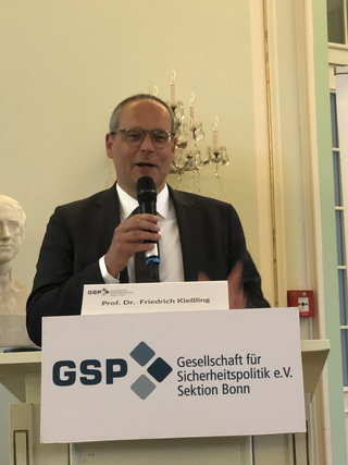 Prof. Dr. Friedrich Kießling spricht an einem Rednerpult mit Mikrofon, ein GSP-Schild ist sichtbar.