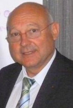 Porträt eines älteren, glatzköpfigen Mannes mit Brille, Anzug und gestreifter Krawatte, leicht lächelnd.