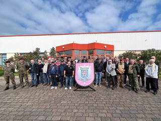 Eine vielfältige Gruppe, darunter Soldaten, posiert mit einem großen rosa Schild mit dem hessischen Wappen vor einem Gebäude.