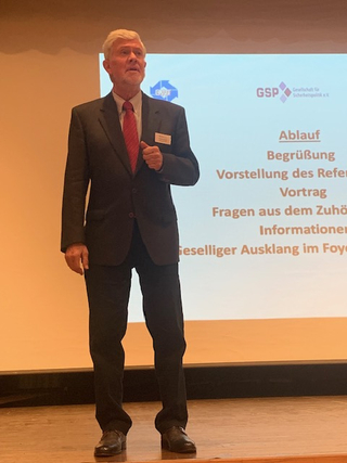 Ein Mann im Anzug und roter Krawatte spricht auf einer Bühne, mit einer Veranstaltungsagenda und GSP-Logo im Hintergrund.