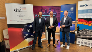Drei Männer stehen lächelnd vor Roll-ups von Organisationen für deutsch-amerikanische Beziehungen und Sicherheitspolitik.