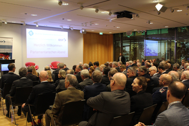 Zuhörer bei einem 'Parlamentarischen Abend' in einem Konferenzraum mit einer Leinwand, die 'Herzlich Willkommen' zeigt.