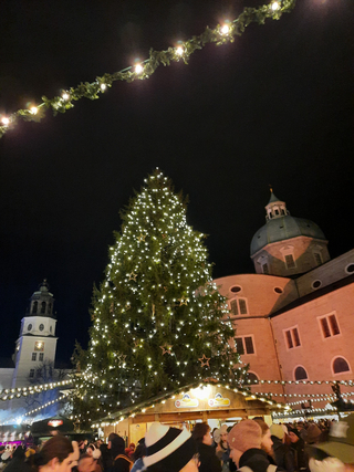 Belebter Weihnachtsmarkt bei Nacht mit großem, beleuchtetem Christbaum, historischen Gebäuden und Lichterketten.