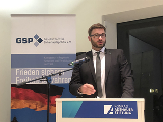 Ein Mann im Anzug mit Brille spricht an einem Rednerpult der Konrad-Adenauer-Stiftung, mit GSP-Banner und deutscher Flagge.