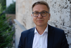 Ein Mann mit kurzen grauen Haaren und Brille in dunklem Anzug und weißem Hemd blickt in die Kamera.