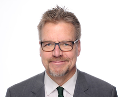 Professionelles Porträt eines lächelnden Mannes mit Brille, kurzen grauen Haaren, Ziegenbart und grauem Anzug mit grüner Krawatte.