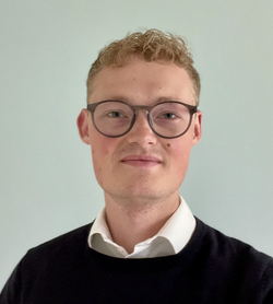 Lachender junger Mann mit lockigem blondem Haar und Brille, dunklem Pullover über weißem Hemd, vor hellblauem Hintergrund.
