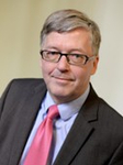 Professionelles Porträt eines lächelnden Mannes mit Brille, grauem Haar, Anzug und rosa Krawatte.