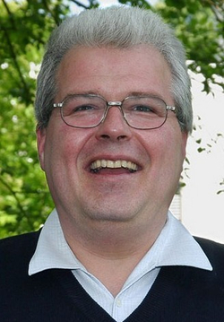 Kopfschuss eines lächelnden Mannes mit grauen Haaren und Brille, der einen dunklen Pullover über einem hellen Hemd im Freien trägt.