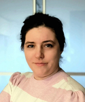 Lächelnde Frau mit dunklen Haaren trägt einen rosa-weiß gestreiften Pullover vor hellblauem Hintergrund.