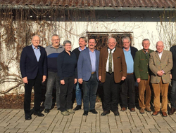 Eine lächelnde Gruppe von zehn Erwachsenen (9 Männer, 1 Frau), leger und teilweise traditionell gekleidet, draußen stehend.