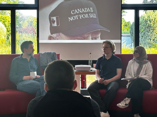 Drei Personen sitzen auf einem roten Sofa und blicken auf die Projektion einer Kappe mit der Aufschrift 'CANADA IS NOT FOR SALE'. Eine Person im Vordergrund.