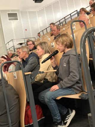 Junge Person mit blonden Haaren hält ein Mikrofon und spricht vor Publikum in einem Hörsaal.